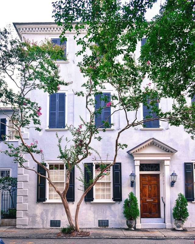 Charleston home with wooden door, classic pediment, pink stucco with black shutters.Pink Crepe myrtles are blooming